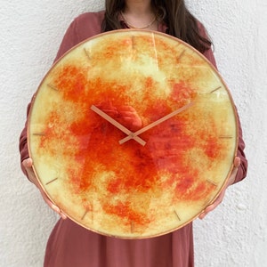 May include: A large, round clock with a yellow and orange marbled face and copper-colored hands. The clock is being held by a person.
