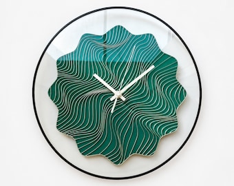 May include: A round wall clock with a thin black frame and a clear face. The center features a scalloped, star-shaped teal insert decorated with intricate, flowing white topographic-style lines. Three slender white hands indicate the time.