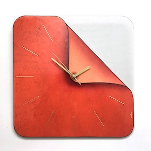 Pode incluir: Um relógio de parede quadrado com um mostrador vermelho e ponteiros dourados. O mostrador do relógio é projetado para parecer uma página sendo virada. O relógio tem um fundo branco atrás do mostrador vermelho.