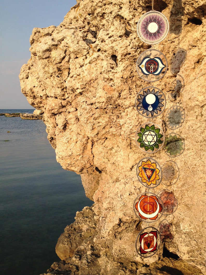 Wall hanging chakras handmade on glass featuring all seven chakra symbols. Spiritual yoga decor and chakra sun catcher set designed for meditation rooms, boho interiors and meaningful handmade gifts for her.
