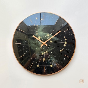 Pode incluir: Um relógio de parede redondo com mostrador preto apresentando uma nebulosa branca e a constelação de Órion em pontos e linhas douradas. O relógio tem ponteiros e moldura na cor cobre. O texto "milli art" está impresso no canto inferior direito.