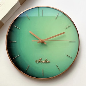 Puede incluir: Reloj de pared redondo con esfera verde degradada y manecillas y marco de color cobre. La esfera del reloj tiene la palabra "Irelia" en cursiva. Un artículo decorativo.