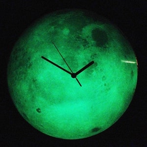 Puede incluir: Un reloj verde que brilla en la oscuridad con forma de luna y agujas negras.