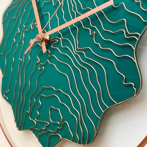 May include: A teal wall clock with a distinctive design. The clock face has a layered, topographic-like pattern in gold against a teal background. The hands are copper-coloured, and the clock is surrounded by a copper-coloured ring.