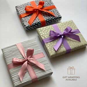 May include: Three square gift boxes wrapped in patterned paper with satin ribbons. One box has black and white dashes with an orange ribbon, one has gold polka dots with a purple ribbon, and one has black stripes with a pink ribbon. Text reads GIFT WRAPPING AVAILABLE.