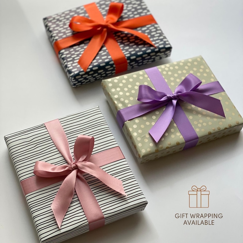 May include: Three square gift boxes wrapped in patterned paper with satin ribbons. One box has black and white dashes with an orange ribbon, one has gold polka dots with a purple ribbon, and one has black stripes with a pink ribbon. Text reads GIFT WRAPPING AVAILABLE.