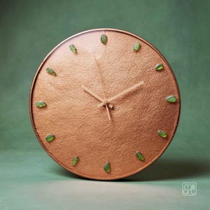 Könnte beinhalten: Eine runde Wanduhr mit einem strukturierten, kupferfarbenen Zifferblatt und einem dünnen, passenden Rahmen. Die Uhr hat zwölf kleine, durchscheinende grüne Meerglasstücke als Stundenmarkierungen und zwei schmale, kupferfarbene Zeiger.