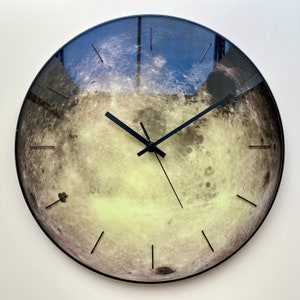May include: A round wall clock with a black frame and black hands. The clock face features a detailed image of the moon's surface.