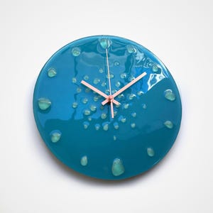 Könnte beinhalten: Eine kreisförmige Wanduhr mit glänzendem blaugrünem Finish und erhabenen, durchscheinenden hellgrünen Tropfen, die über die Oberfläche verteilt sind. Die Uhr hat drei dünne, metallisch roségoldene Zeiger, die mittig auf dem Zifferblatt angebracht sind.