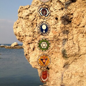 Set of 7 chakra glass suncatchers hanging outdoors near coastal rocks. Handpainted mandala glass wall hanging chakras decor for mindfulness spaces, yoga studios, meditation rooms and spiritual gifts.