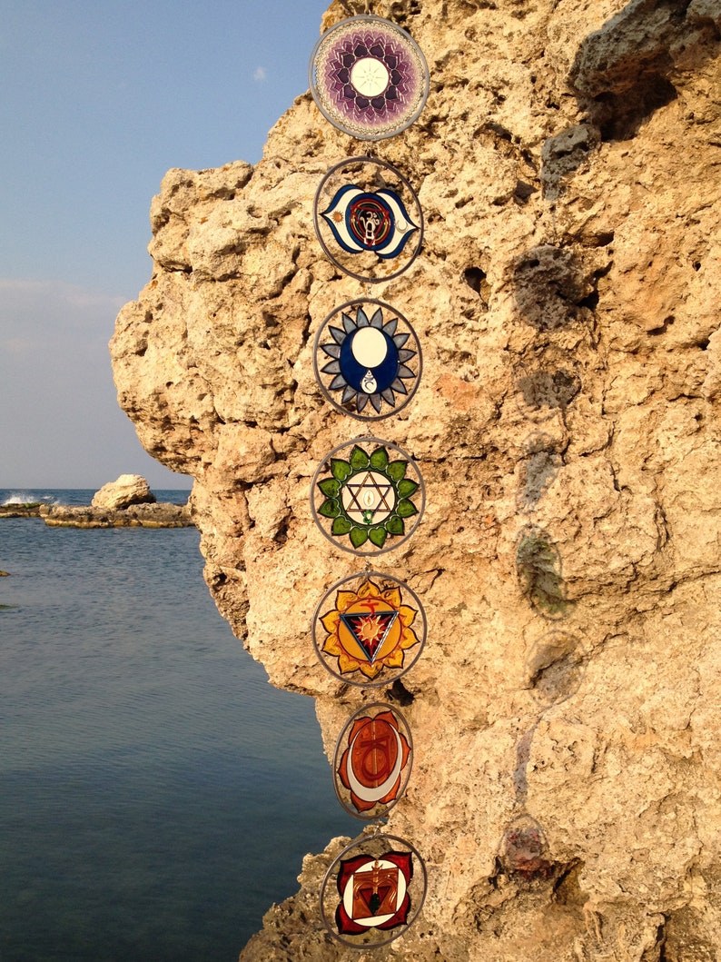 Set of 7 chakra glass suncatchers hanging outdoors near coastal rocks. Handpainted mandala glass wall hanging chakras decor for mindfulness spaces, yoga studios, meditation rooms and spiritual gifts.