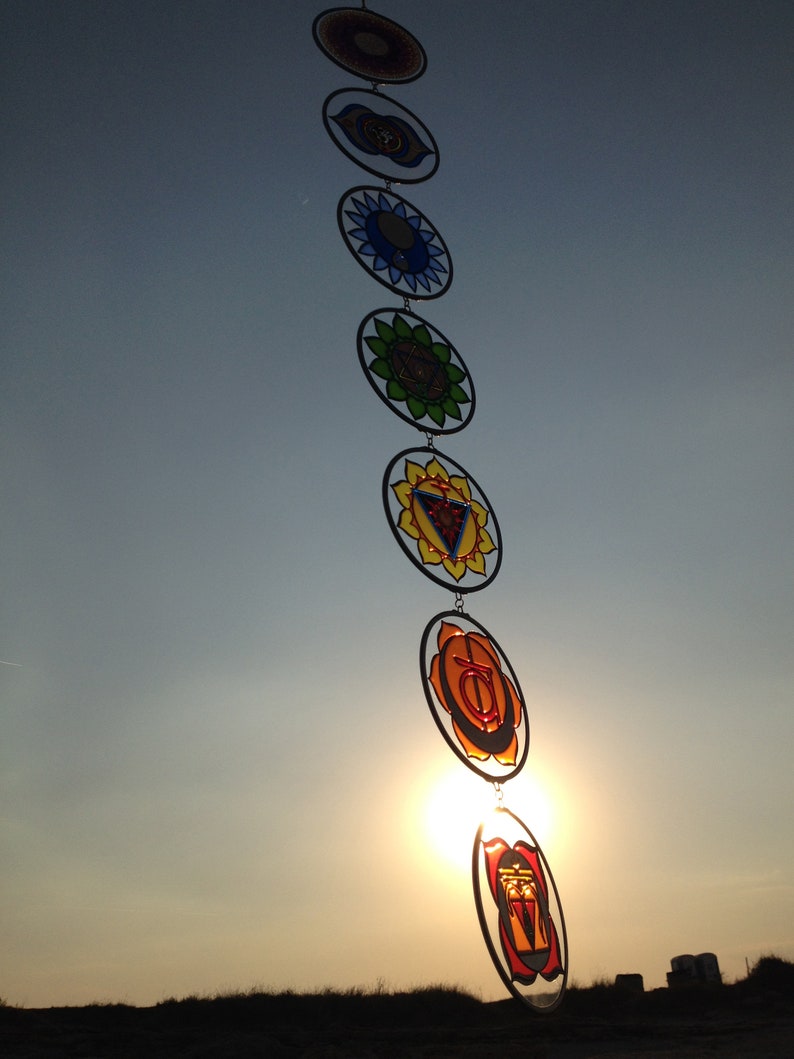 Seven chakra mandala glass suncatchers glowing beautifully in sunlight during sunset. Handmade wall hanging chakras decor for yoga room, meditation space, spiritual home styling and chakra balancing energy.