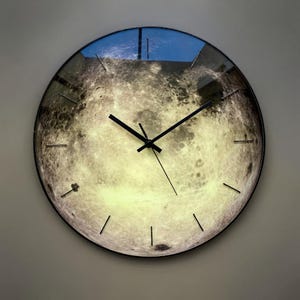 Op de afbeelding: Ronde wandklok met een gedetailleerde afbeelding van het maanoppervlak. De klok heeft een zwart frame, zwarte wijzers en eenvoudige zwarte uurmarkeringen. De maanfoto heeft een gloeiende, getextureerde uitstraling, met lichte en donkere gebieden.