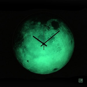 May include: A circular wall clock featuring a glowing green lunar surface design with detailed craters and textures. The clock has three thin black hands positioned at the centre, set against a dark background. The text milli art is visible.