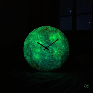 Mercury wall clock glowing green in a dark room after charging with light, showing the luminous phosphorescent details across the planetary surface.