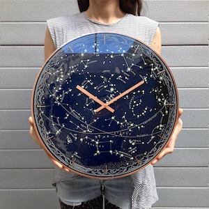 Könnte beinhalten: Eine runde Wanduhr mit einem tiefblauen Zifferblatt, das eine detaillierte weiße Sternenkarte und Sternbildillustrationen zeigt. Die Uhr hat einen dünnen kupferfarbenen Rahmen sowie passende kupferfarbene Stunden- und Minutenzeiger.