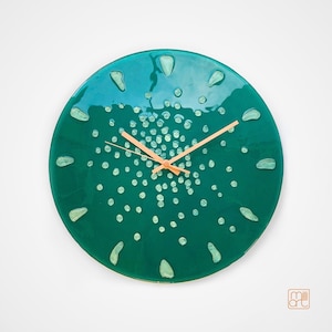 Op de afbeelding: Een ronde wandklok met een glanzend blauwgroen oppervlak, voorzien van een centraal cluster van kleine lichtgroene stippen en twaalf grotere druppelvormige markeringen langs de rand. De klok heeft dunne, lichte wijzers en de tekst milli art.