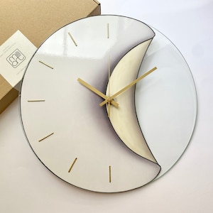 May include: A white and grey glass wall clock with gold hands and a crescent moon design. The clock has a clear glass outer ring.