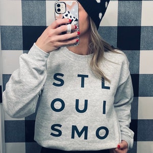 May include: A gray sweatshirt with the text "STL SOUI SMO" printed on the front. The person is wearing black pants and a black and white polka dot beanie.