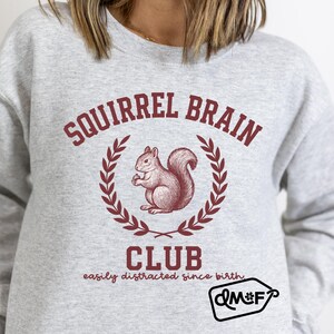 May include: A light gray sweatshirt with the text "SQUIRREL BRAIN CLUB" in maroon. A maroon illustration of a squirrel holding a nut is in the center, surrounded by a laurel wreath. Below the club name is the text "easily distracted since birth."