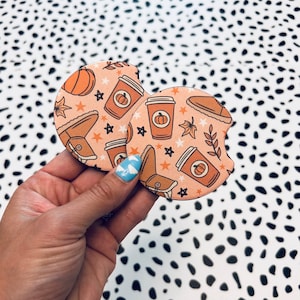 May include: Two round, peach-colored drink coasters with a fall-themed design. The coasters feature illustrations of coffee cups, boots, pumpkins, and stars.