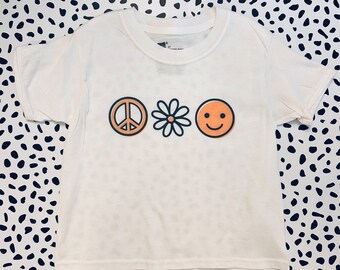 May include: White t-shirt with a peace sign, daisy, and smiley face graphic in orange.