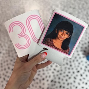 Pode incluir: Dois suportes de lata brancos. Um tem o número "30" em rosa, o outro tem uma foto de uma pessoa com cabelo longo e preto. Ambos os suportes têm uma borda rosa. Os suportes são segurados por uma mão com manicure de ponta vermelha.