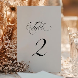May include: A table setting with a white card displaying "Table 2" in elegant black script. The scene includes a lit candle, clear glass, and dried floral arrangements, suggesting a wedding or event.
