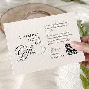 May include: A cream-colored card with elegant script reading "A Simple Note on Gifts." The card includes a QR code and text about a honeymoon fund. The card is held over a white surface with greenery and wine glasses in the background.