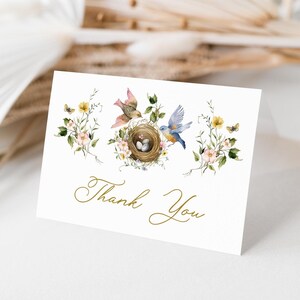 Pode incluir: Um cartão de agradecimento branco com uma ilustração em aquarela de dois pássaros em um ninho com ovos. O ninho é cercado por flores e folhas. O texto "Obrigado" está escrito em letras douradas.
