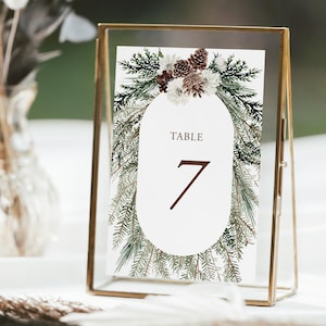 May include: A table number card in a gold frame. The card features a white oval with the word "TABLE" and the number "7" in brown. The oval is surrounded by green and brown floral elements.