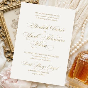Op de afbeelding: Een trouwuitnodiging met gouden kalligrafie, een sierlijke gouden lijst, een parelketting, een parfumflesje en een vintage broche. De uitnodigingstekst luidt "Elizabeth Davies to Jacob Alexander Wilson". De scène speelt zich af op een witte kanten achtergrond.