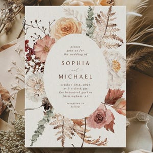 Puede incluir: Invitación de boda con ilustraciones florales en tonos melocotón, burdeos y crema. La invitación incluye los nombres "Sophia" y "Michael", junto con la fecha y hora de la boda. Flores secas y sobres rodean la invitación.
