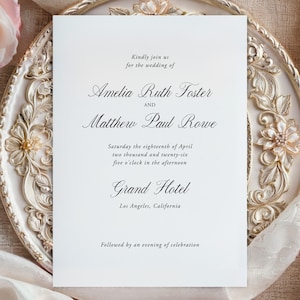 May include: A wedding invitation on a decorative, ornate plate with floral accents. The invitation is white with elegant black script, including the names Amelia Ruth Foster and Matthew Paul Rowe, and the Grand Hotel in Los Angeles.