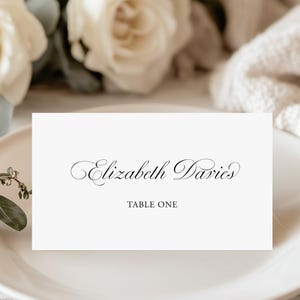 Op de afbeelding: Een witte naamkaart met de naam "Elizabeth Davies" in elegant schrift en "TABLE ONE" eronder gedrukt. De kaart ligt op een crèmekleurige plaat, met witte rozen en groen op de achtergrond.