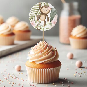 May include: A cupcake with pink and white frosting and sprinkles topped with a round cake topper featuring a cartoon monkey hanging from a vine with pink flowers and green leaves.