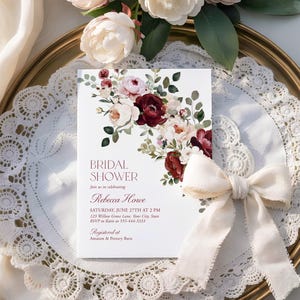 May include: A white rectangular bridal shower invitation card featuring a floral arrangement of deep burgundy and soft pink roses with green leaves. The text reads: BRIDAL SHOWER, Join us in celebrating, Rebecca Howe, SATURDAY, JUNE 27TH AT 2 PM.
