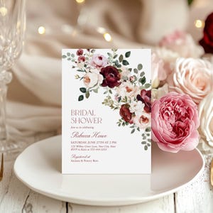 May include: A rectangular bridal shower invitation card featuring a watercolor floral arrangement of deep burgundy, blush pink, and cream roses with green eucalyptus leaves. The text reads: BRIDAL SHOWER, join us in celebrating, Rebecca Howe, SATURDAY, JUNE 27TH AT 2 PM, 123 Willow Grove Lane, Your City, State, RSVP to Katie at 555-444-3333, Registered at Amazon & Pottery Barn.