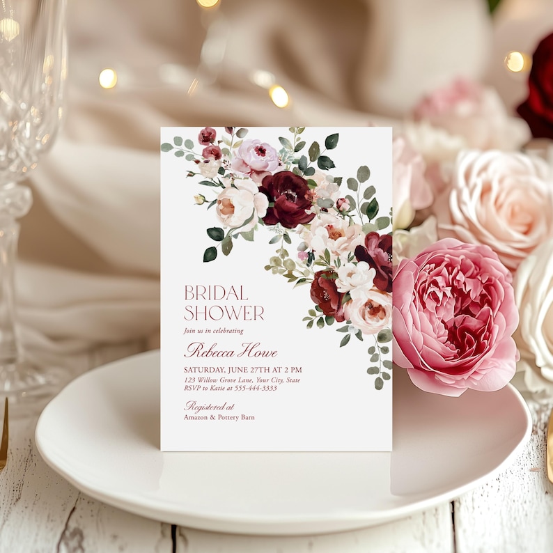 May include: A rectangular bridal shower invitation card featuring a watercolor floral arrangement of deep burgundy, blush pink, and cream roses with green eucalyptus leaves. The text reads: BRIDAL SHOWER, join us in celebrating, Rebecca Howe, SATURDAY, JUNE 27TH AT 2 PM, 123 Willow Grove Lane, Your City, State, RSVP to Katie at 555-444-3333, Registered at Amazon & Pottery Barn.