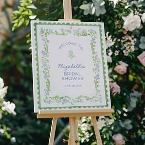 K&ouml;nnte beinhalten: Ein wei&szlig;es Schild auf einer Holzstaffelei mit einem gr&uuml;nen, gewellten Rand und floralen Akzenten. Auf dem Schild steht "Willkommen zu Elizabeths Bridal Shower" mit dem Datum 28. Juni 2025. Der Hintergrund zeigt Gr&uuml;n und Blumen.