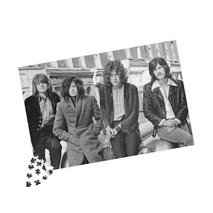 May include: Black and white photograph of the rock band Led Zeppelin. The four band members are dressed in vintage clothing, posing in front of a building and a car. The image is a puzzle piece.