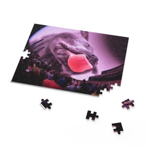 May include: A jigsaw puzzle featuring a close-up of a gray animal with a pink tongue extended. The background shows a crowd of people in a stadium setting, with a purple and pink color scheme.