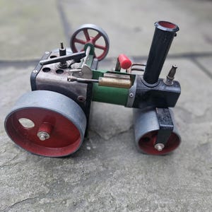 Puede incluir: Un modelo de apisonadora a vapor en miniatura que presenta una caldera cilíndrica verde, una chimenea negra y grandes ruedas grises con centros rojos. La construcción metálica incluye una pequeña manija roja, tuberías de latón y un volante.
