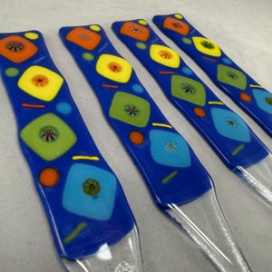 May include: Four blue glass serving utensils with colorful geometric designs. Each piece features a clear handle and a vibrant blue top adorned with squares, circles, and diamond shapes in orange, yellow, green, and blue.