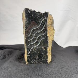 May include: A carved stone sculpture featuring a dark, speckled surface with a light-colored, wave-like design. The edges of the stone are rough and tan. The sculpture is standing upright.