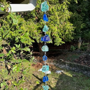 May include: A blue and green glass wind chime hanging from a metal hook. The chime is made of various sized pieces of glass, some of which are clear and some of which are blue or green. The chime is hanging in a garden setting.