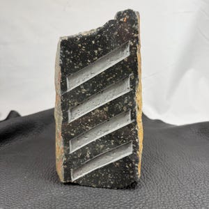 May include: A rectangular, dark gray stone with a rough, textured surface. The stone features a series of diagonal, parallel grooves carved into its face. The grooves are a lighter gray color, creating a contrast against the darker stone.