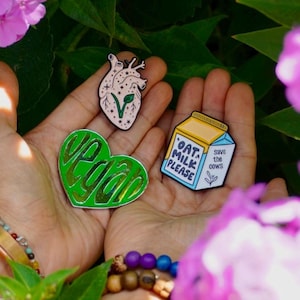 Op de afbeelding: Drie emaillepins met een veganistisch thema. De pins hebben de vorm van een hart, een pak havermelk en een hart met een plant die eruit groeit. De hartpin zegt "vegan" in groene letters. Het pak havermelk zegt "Havermelk alstublieft" en "Red de koeien".