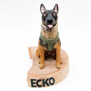 May include: A hand-crafted ceramic figurine of a German Shepherd dog wearing a police vest and a collar with the word "POLICE" on it. The dog is sitting on a wooden base with the name "ECKO" written on it.