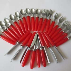 Puede incluir: Un conjunto de cubiertos en miniatura con mangos rojos y utensilios de metal plateado. El juego incluye cucharas, tenedores y cuchillos, dispuestos en un patrón circular. Los mangos son de un rojo vibrante.